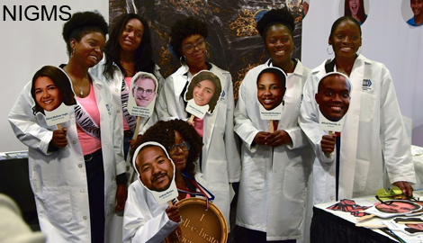 Six women in lab coats, each holding up a photo of another person's face